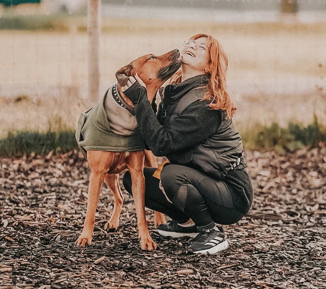 Frau hockt auf dem Trainingsplatz, ihr brauner Hund schmiegt sich an ihr Gesicht